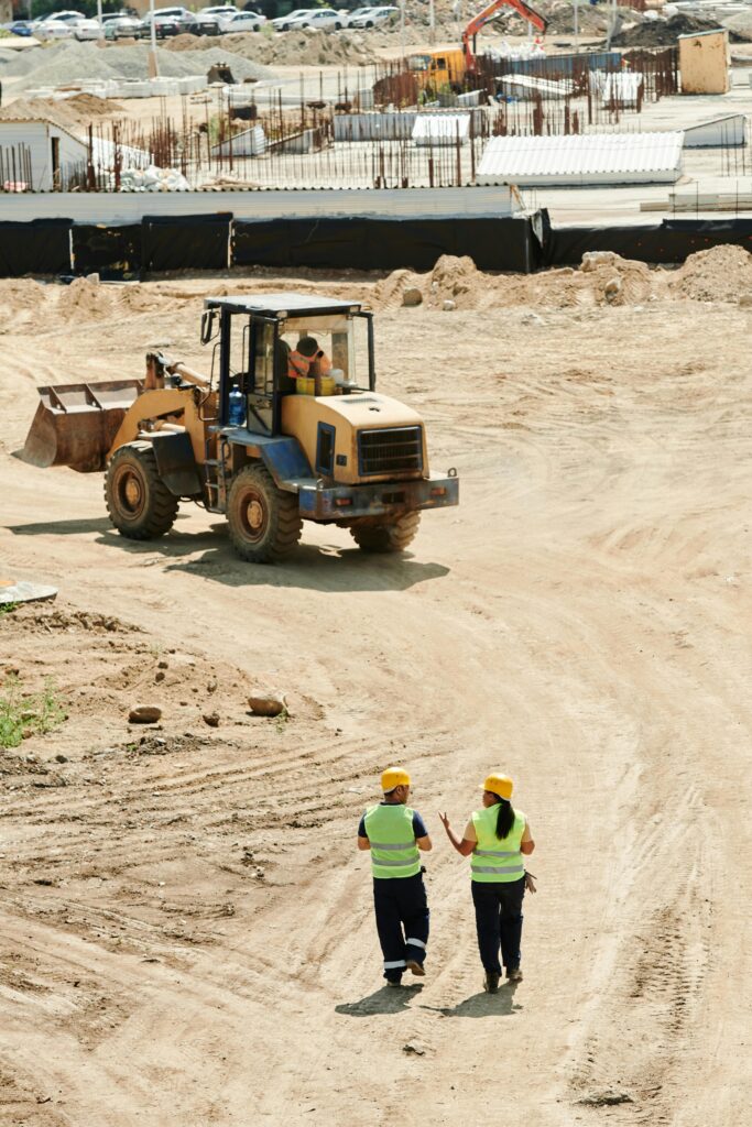 Construction workers in safety gear walking at a busy site with a backhoe in operation.